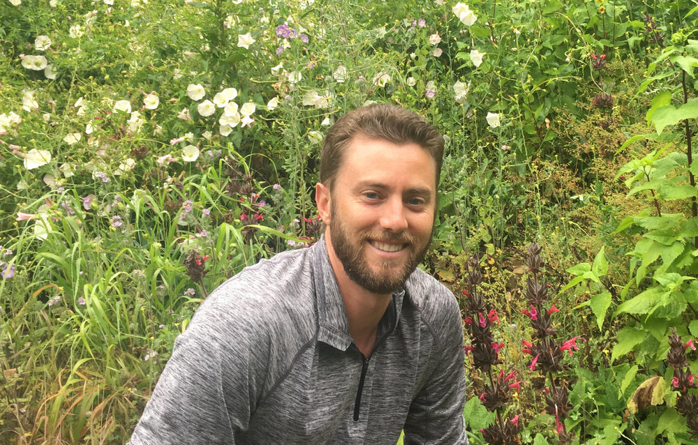 Man with a beard kneeling down among tall grass and flowers